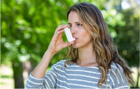 Persoon in een park gebruikt een inhalator, omringd door groen, gekleed in een grijs-wit gestreept shirt.
