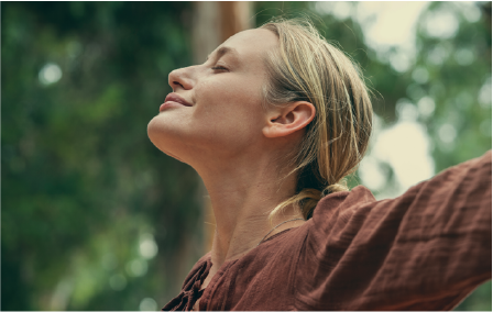 Persoon met gesloten ogen en gestrekte armen in een groene buitenomgeving, straalt rust en verbondenheid met de natuur uit.