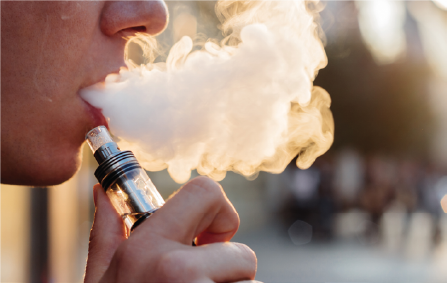 Close-up van een persoon die een vape gebruikt en een grote dampwolk uitblaast, met een metallic vape-apparaat en een wazige buitenachtergrond.